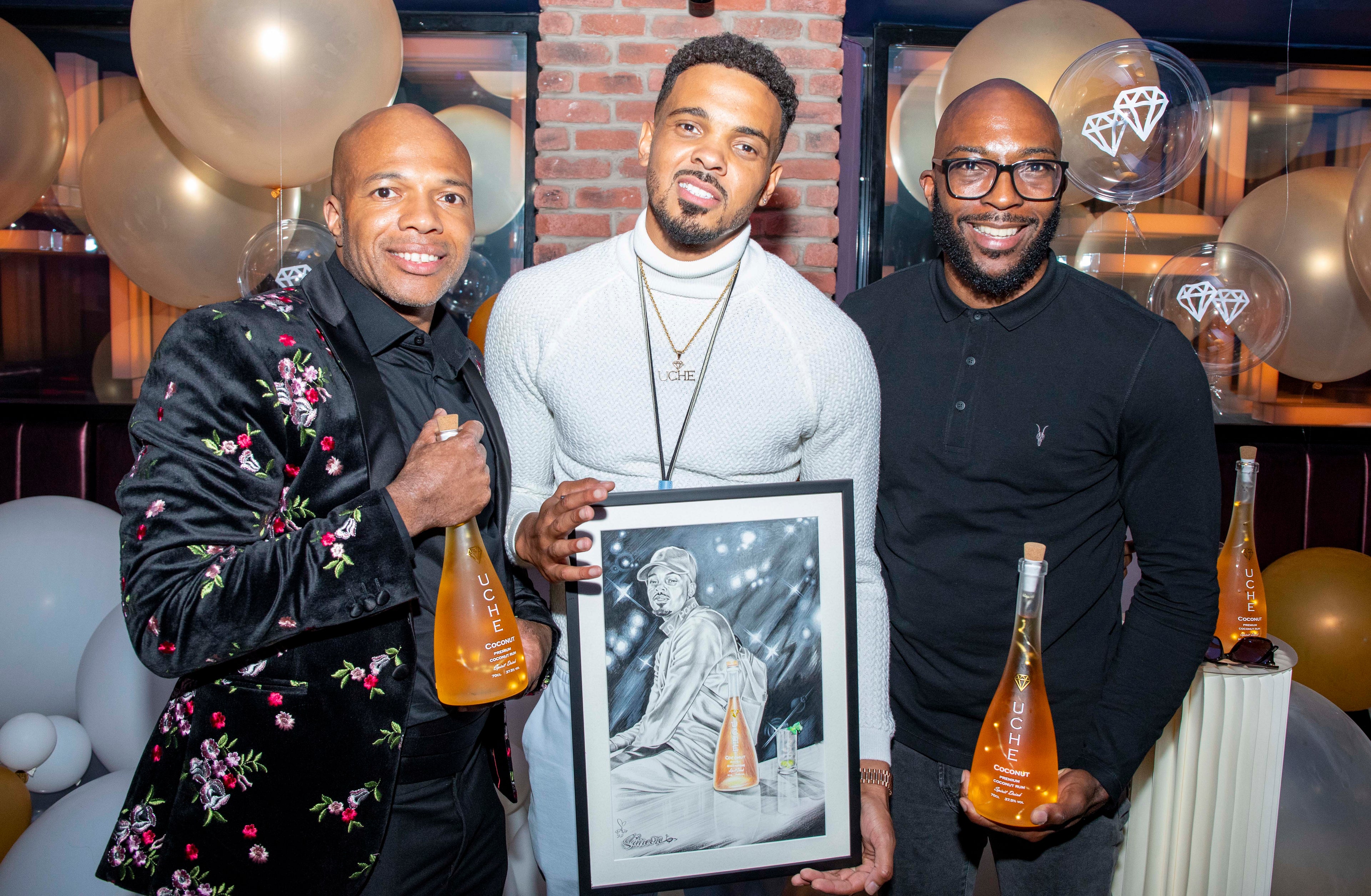 Three men posing with a framed picture and bottles at an event with balloons and decor.
